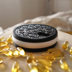 Op de afbeelding: Een zwart-wit Oreo-koekjesvormige container met het woord "OREO" erop. De container heeft een witte vulling en is omgeven door gele capsules, waarschijnlijk vitamines of supplementen. De container staat op een houten oppervlak.