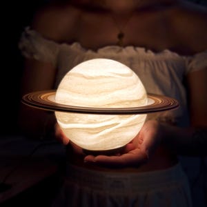 Może przedstawiać: Świecąca lampa w kształcie Saturna z szczegółowym systemem pierścieni. Lampa emituje miękkie, ciepłe światło, ukazując wirujące wzory powierzchni planety. Tło jest ciemne.