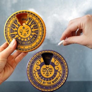 Pode incluir: Dois organizadores de comprimidos circulares com desenhos de sol e lua. Um é amarelo com o texto "LIVE BY THE SUN", o outro é roxo com o texto "LOVE BY THE MOON". Uma mão segura um comprimido branco acima do organizador roxo.