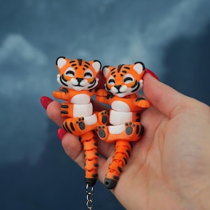 Peut inclure: Deux porte-clés tigres orange et blancs avec des rayures noires et des visages souriants. Les porte-clés sont tenus dans une main sur un fond bleu. Chaque porte-clés a une chaîne et un anneau argentés.