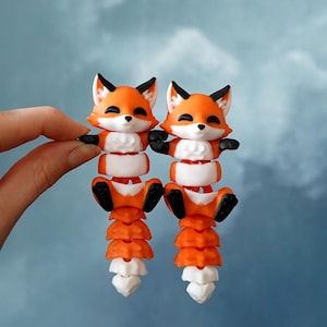 May include: Two whimsical fox figurines, each with orange and white segmented bodies, black paws, and pointed ears. The foxes have smiling faces and are set against a cloudy blue background. The figurines are held by a hand.