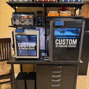Peut inclure: Installation d'un service d'impression 3D personnalisé avec deux imprimantes 3D, des bobines de filament et des unités de stockage. Les imprimantes affichent le texte "CUSTOM 3D PRINTING SERVICE". L'installation comprend des étagères noires, un bureau noir et un meuble gris foncé à tiroirs.