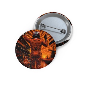 Op de afbeelding: Een ronde button met een speld aan de achterkant, voorzien van een afbeelding van een autocoureur in een oranje pak met opgeheven armen. De tekst luidt DUTCH DON'T GIVE A FUCK MENTALITY. De achtergrond toont een zonsondergang, windmolens en een circuit.