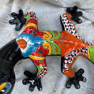 May include: A brightly painted ceramic lizard sculpture. The lizard has a glossy black body and is decorated with a floral design in blue, orange, green, and red. The feet are black. This decorative piece is ideal for adding a touch of art to any space.