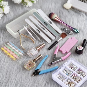 May include: A selection of manicure tools and accessories, including nail files, brushes, clippers, nail varnish, and a container of rhinestones. The items are arranged on a soft, grey surface, suggesting a beauty or manicure theme.