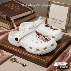 K&ouml;nnte beinhalten: Wei&szlig;e Minnie Mouse Kinderclogs mit einem Minnie Mouse Cartoon-Motiv. Die Clogs haben einen Riemen und werden auf einem Holztablett pr&auml;sentiert. Der Text "Minnie Mouse Kids Slippers" ist sichtbar.