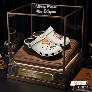 K&ouml;nnte beinhalten: Wei&szlig;e und schwarze Mickey Mouse Kinderslipper in einer Glasvitrine. Die Slipper zeigen eine Cartoon-Grafik von Mickey Mouse und Minnie Mouse. Die Vitrine hat einen goldenen Rahmen und einen Marmorsockel.