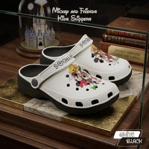 Op de afbeelding: Witte en zwarte Mickey and Friends kinderslippers. De slippers hebben een cartoonafbeelding van Mickey Mouse en zijn vrienden. De tekst "Mickey and Friends Kids Slippers" is zichtbaar. De slippers worden op een marmeren oppervlak gepresenteerd.