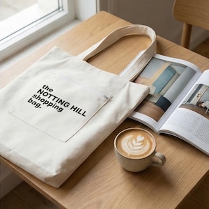 Puede incluir: Un bolso tote de color crema con el texto "the NOTTING HILL shopping bag" en negro. Una taza de café y una revista abierta están sobre una mesa de madera.