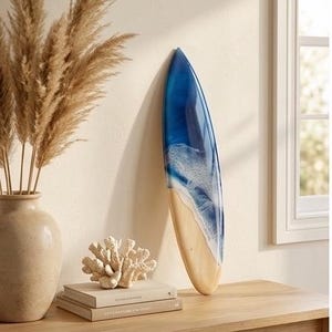 May include: A decorative surfboard wall art piece featuring a smooth wooden base transitioning into a glossy blue resin finish with white foam detailing that mimics ocean waves crashing against a sandy shore, displayed in a vertical orientation.