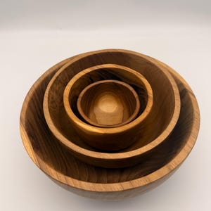 Peut inclure: Ensemble de quatre bols en bois emboîtés. Les bols sont en bois brun clair avec des motifs de grain visibles. Les tailles varient, le plus grand en bas et le plus petit au centre.