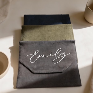 May include: Three fabric pouches in shades of gray, olive green, and navy blue are stacked. The front pouch is gray and features the name "Emily" in white script. A beige mug and bowl are visible in the background.