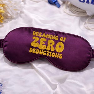 May include: A deep purple satin sleep mask with the text "Dreaming of Zero Deductions" in gold glitter lettering. The mask has an elastic band and is designed to block out light for better sleep.