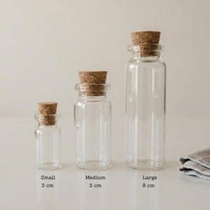 Può includere: Tre bottiglie di vetro trasparente con tappi di sughero, etichettate "Small" (3 cm), "Medium" (5 cm) e "Large" (8 cm). Le bottiglie sono vuote e si trovano su una superficie bianca. Un panno grigio piegato è nell'angolo in basso a destra.