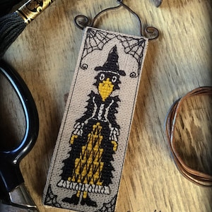 May include: A cross-stitch wall hanging featuring a black and yellow witch with a black hat and a spiderweb design. The text "Copyright 2020 www.TheElfinForest.etsy.com" is at the bottom of the hanging.