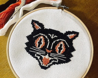 May include: A black and orange cross-stitch embroidery of a cat's face in a wooden embroidery hoop. The cat has large, round eyes and a wide, toothy grin.