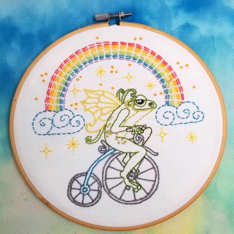 A pre-printed cotton fabric embroidery design stamped onto a 10.5" x 10.5" white fabric panel. Features a frog fairy riding a bicycle through the clouds with a rainbow and starbursts.