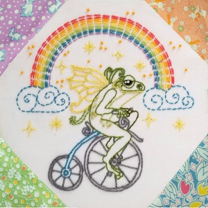A hand embroidery design featuring a frog fairy riding a bicycle and a rainbow.  Quilted display idea. Pre-printed stamped pattern for you to stitch along the lines.