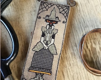 Pode incluir: Um padrão de ponto cruz de um gato preto vestido de bruxa com um chapéu pontudo e uma vassoura. O gato está segurando um caldeirão e está rodeado por um desenho de teia de aranha. O padrão está em um tecido bege.