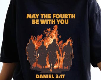 May include: Black t-shirt featuring the gold text "MAY THE FOURTH BE WITH YOU". Below the text is an illustration of four figures walking towards a fire. The text "DANIEL 3:17" is printed at the bottom of the shirt.