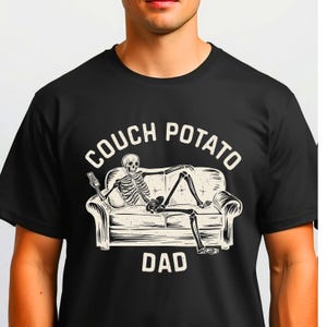 May include: Black t-shirt with a graphic of a skeleton lounging on a couch, holding a remote. The words "COUCH POTATO DAD" are printed above and below the image in a white, bold font.