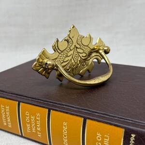 May include: A vintage brass drawer pull with an ornate leaf design, resting on a brown book. The book has a gold spine with the text "READER'S DIGEST CONDENSED BOOKS" and other titles. The drawer pull is a warm gold color.
