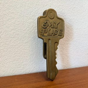 May include: A brass-colored metal door knocker shaped like a large key. The head of the key features the embossed text MY PLACE in a stylized font. The object has a vertical orientation with a hinged mechanism at the top for the knocking action.