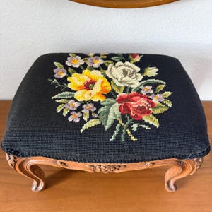 May include: A small, square footstool with a black needlepoint top featuring a floral design in yellow, red, white, and purple. The wooden legs are carved with ornate details and have a curved shape. The footstool is a decorative accent piece.