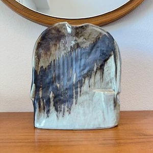 May include: A flat, arched ceramic vase with a glossy finish. The surface features a cream-colored base with dark brown and black glaze dripping vertically from the top edge. The piece has a small, rectangular opening on the lower right side.