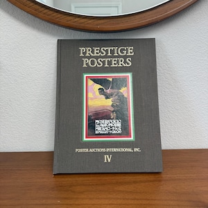 May include: A hardcover book titled "PRESTIGE POSTERS" with a colorful image of a vintage poster. The book's cover is dark gray with gold lettering and a framed poster image. The book is on a wooden surface.