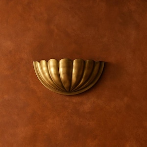 May include: A decorative, half-circle wall sconce in a warm, antique gold finish. The sconce features a scalloped design, with vertical ridges creating a textured surface. It is mounted on a textured, reddish-brown wall.