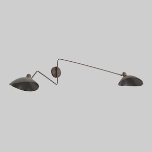 May include: A bronze-toned wall sconce with two lights, showcasing a minimalist design. It has two curved, dome-shaped shades and a central circular mounting plate. The arms extend horizontally, offering a balanced, contemporary look.