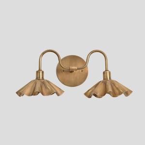 Puede incluir: Aplique de pared en tono latón con dos luces. Cada luz tiene una pantalla con volantes y un brazo curvo. El accesorio está montado en una placa trasera circular, creando una estética vintage.