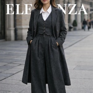 May include: A charcoal gray three-piece suit with a long coat. The suit includes a long coat, a vest, and wide-leg trousers. A white collared shirt is worn underneath the vest. The word "ELEGANZA" is printed at the top of the image.