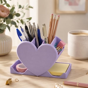 May include: A lavender heart-shaped desk organizer holds pens, pencils, and other stationery items. The organizer has a tray for sticky notes and tape. A cup of coffee and a vase of flowers are in the background.