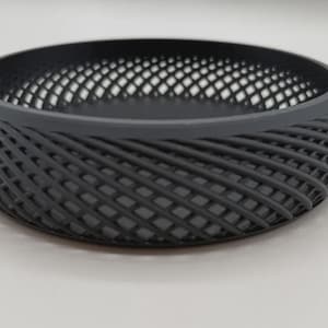 Può includere: Una ciotola rotonda grigio scuro con un design a griglia. La ciotola ha una base piatta e una parte superiore ampia e aperta. I lati sono costituiti da linee intrecciate, creando una superficie strutturata. La ciotola è probabilmente realizzata in plastica o materiale simile.