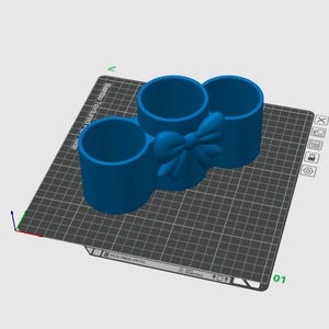 May include: A blue 3D-printed desk organizer with three cylindrical compartments. A bow detail is centered between the compartments. The organizer is on a gray grid surface.