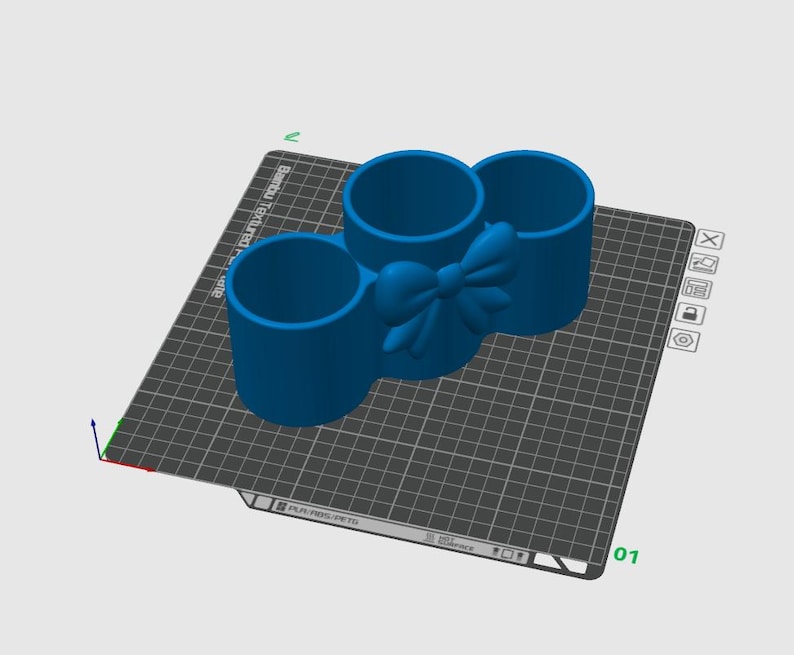 May include: A blue 3D-printed desk organizer with three cylindrical compartments. A bow detail is centered between the compartments. The organizer is on a gray grid surface.