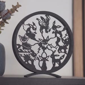 May include: A circular black wooden cutout featuring a central Eevee surrounded by its eight evolutions in a radial pattern. The intricate laser-cut design displays silhouettes of Vaporeon, Jolteon, Flareon, Espeon, Umbreon, Leafeon, Glaceon, and Sylveon.