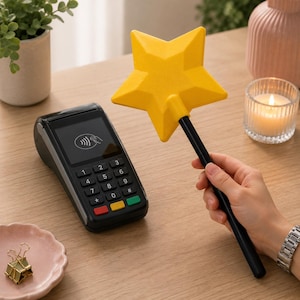 May include: A yellow star-shaped wand with a black handle is held near a black point-of-sale (POS) terminal. The terminal has a screen displaying a contactless payment symbol and a keypad with coloured buttons. A lit candle and a small plant are in the background.