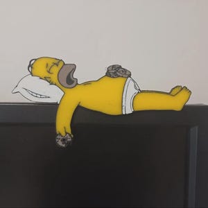 May include: A yellow cartoon character figure with a brown beard, wearing white underwear, sleeping on a white pillow. The figure has a stack of brown circular treats on its stomach and is holding another brown circular treat in its right hand.