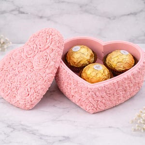 Puede incluir: Una caja rosa en forma de corazón con un patrón de rosas, que contiene tres bombones envueltos en oro. La caja está abierta, con la tapa al lado. El fondo es una superficie de mármol.