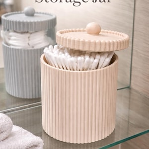 May include: A ribbed storage jar with a lid, filled with cotton swabs. The jar is light peach, with a matching lid and a small wooden knob. The text "Ribbed Storage Jar" is at the top. The text "STL File" is at the bottom.