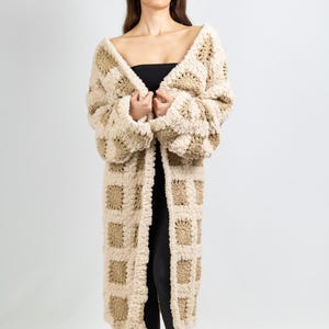 May include: A long, open-front cardigan in a checkerboard pattern. The beige and tan knitwear has a soft, textured appearance. The cardigan is floor-length and has long sleeves. The model is wearing a black top and leggings.