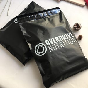 Könnte beinhalten: Zwei schwarze Poly-Versandtaschen mit dem Text "OVERDRIVE NUTRITION" in Weiß. Die Taschen haben eine glänzende Oberfläche und eine versiegelte Oberseite. Ein weißes Logo ist ebenfalls sichtbar. Der Hintergrund ist hell.