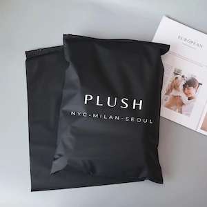 Op de afbeelding: Twee zwarte verzendzakken met het woord "PLUSH" in het wit en de steden NYC, Milaan en Seoul eronder gedrukt. Op de achtergrond is een tijdschrift met het woord "EUROPEAN" te zien.