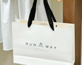 May include: A white shopping bag with black handles, featuring the text "RUN AWAY Femmes" in black. The bag is held by a wooden hand. The background includes a white garment and a wooden surface.