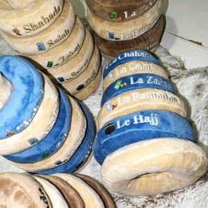 Könnte beinhalten: Eine Sammlung von plüschigen, donut-förmigen Kissen in Beige, Blau und Braun. Jedes Kissen ist mit weißem Text und kleinen Illustrationen bestickt. Der Text enthält Wörter wie "Shahada", "Salah" und "Zakat".