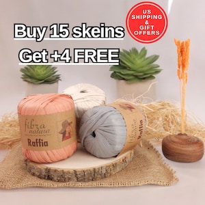 May include: Three skeins of raffia yarn in peach, cream, and gray, displayed on a wooden slice. The peach skein has a label that reads "fibra natura Raffia". The image also includes text that says "Buy 15 skeins Get +4 FREE".