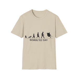 Puede incluir: Camiseta beige con un gráfico negro que representa la evolución de un esquiador, desde la infancia hasta el esquí. El texto "BORN TO SKI" está impreso debajo del gráfico.
